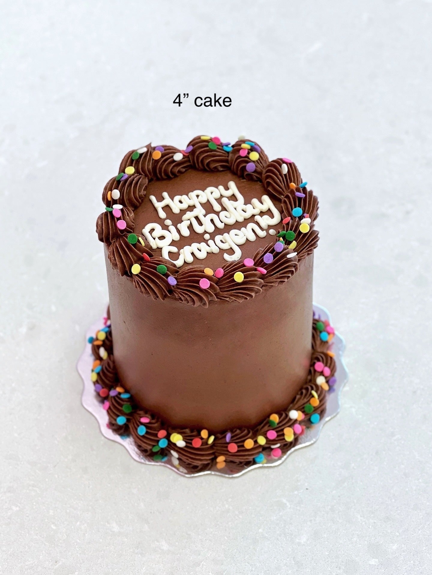 Chocolate birthday cake with colorful sprinkles and 'Happy Birthday!' text on a white background.